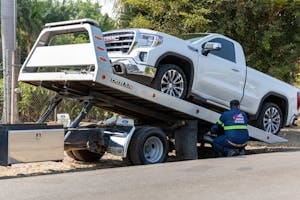 Tow truck operator loading white GMC pickup truck on street in daytime.